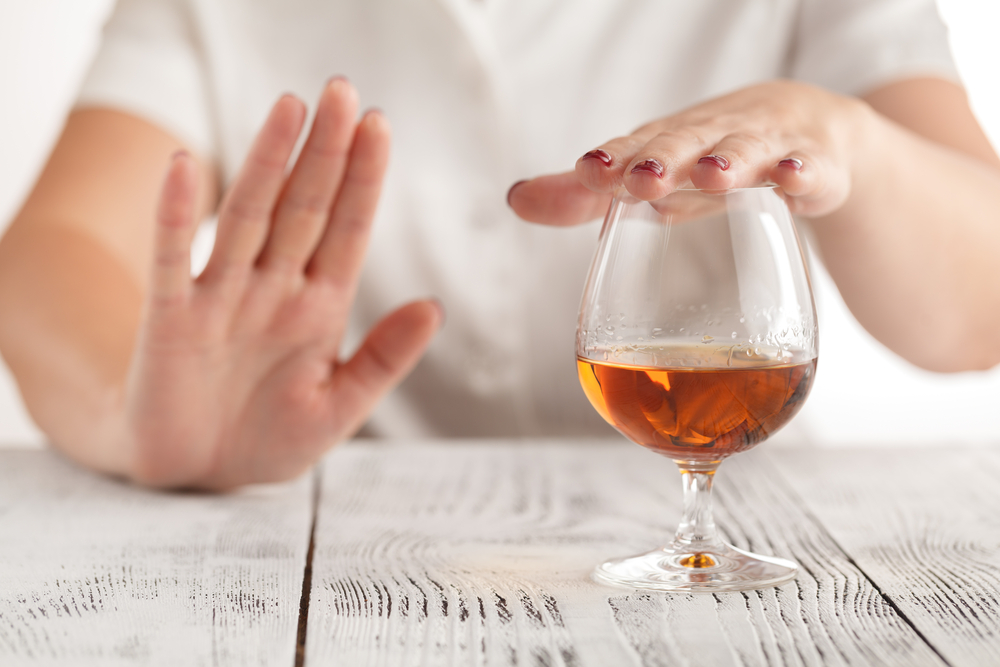 At chest level white female wearing white shirt sitting at table one hand covering top of whiskey glass while other hand held out in the halt position.