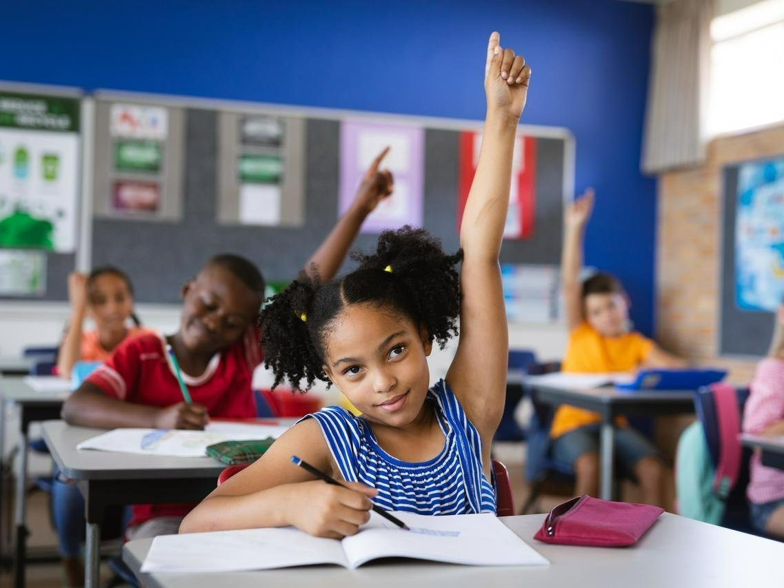 young african american female with pigtails sits at desk in classroom with students and she raises her hand